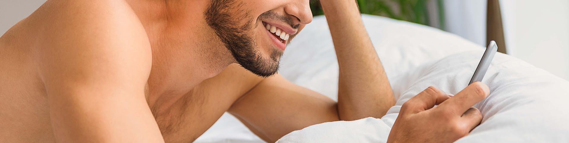 A bearded man smiling and laying down on a bed while looking at his phone.