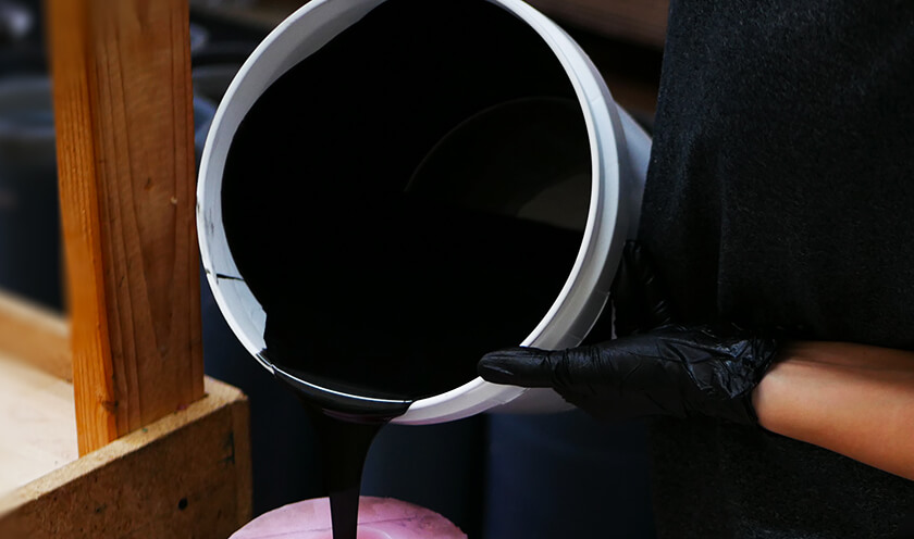 A person with black gloves and a bucket pouring black paint onto a product.