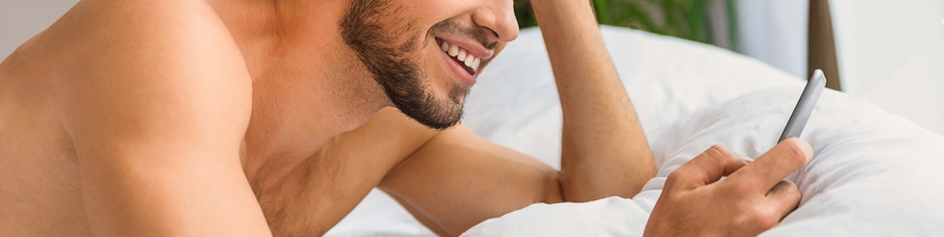 A shirtless man with a beard laying on their side on a bed, smiling and holding a cell phone.